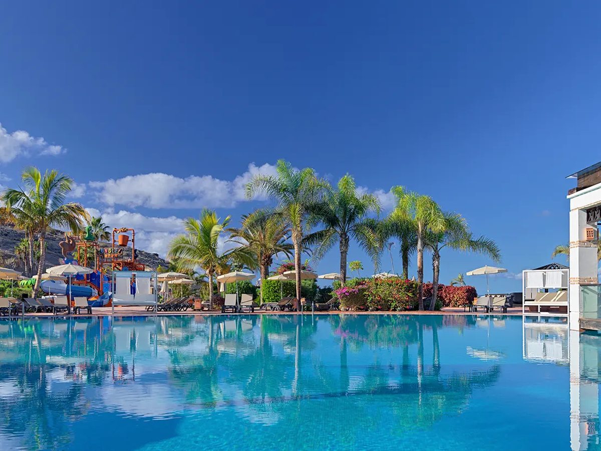 A large free-form outdoor pool reflecting tall palm trees, edged with sun loungers and parasols under a clear blue sky, with the Jungle Park water structure beyond.