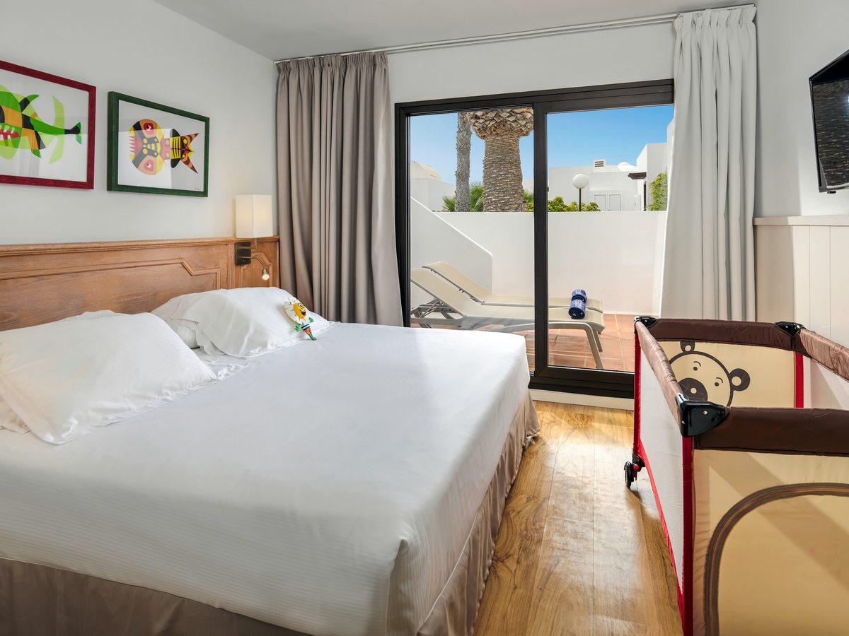 A suite bedroom with a large double bed, a wooden headboard and framed fish prints, a travel cot beside it and sliding doors onto a terrace with a sun lounger.