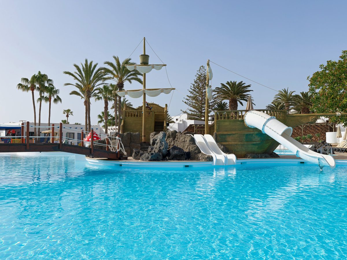 A wooden pirate ship at the poolside with two white open slides and an enclosed white tube slide running down into clear turquoise water.