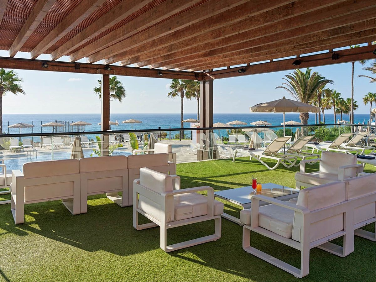 Covered pergola terrace with white cushioned sofas and armchairs on artificial grass, looking past palms to the pool and the sea.