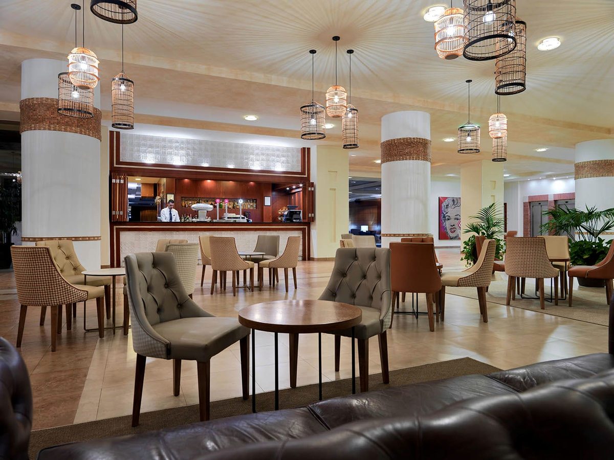 Lobby lounge bar with a staffed counter, wicker-cage pendant lights, cream columns and clusters of armchairs around low tables.