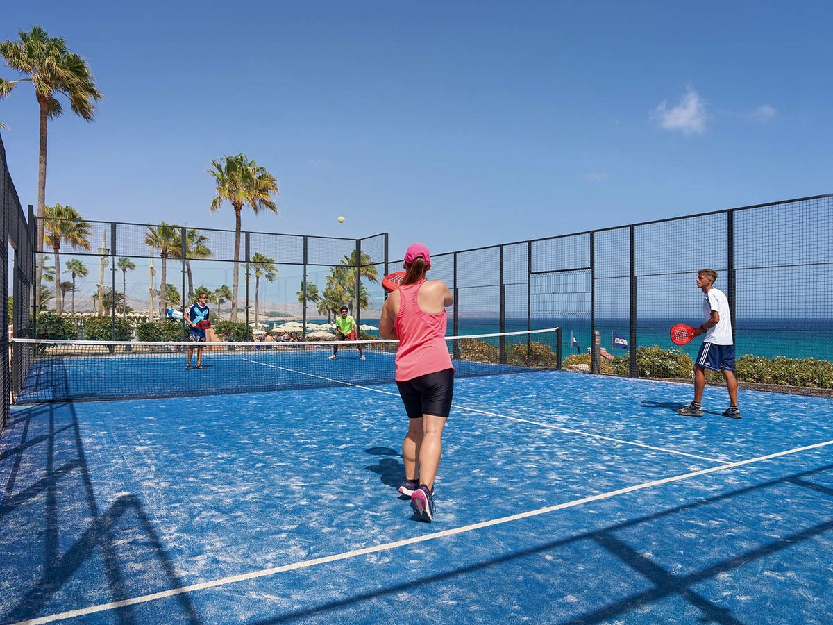 Two players on a blue-surfaced padel court enclosed by glass and mesh, with palms and the sea behind.