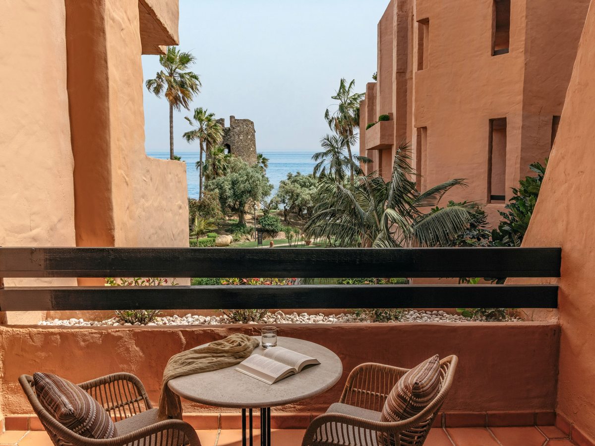 A terracotta room balcony with wicker chairs and a small table, looking past palms to a stone watchtower and the sea.