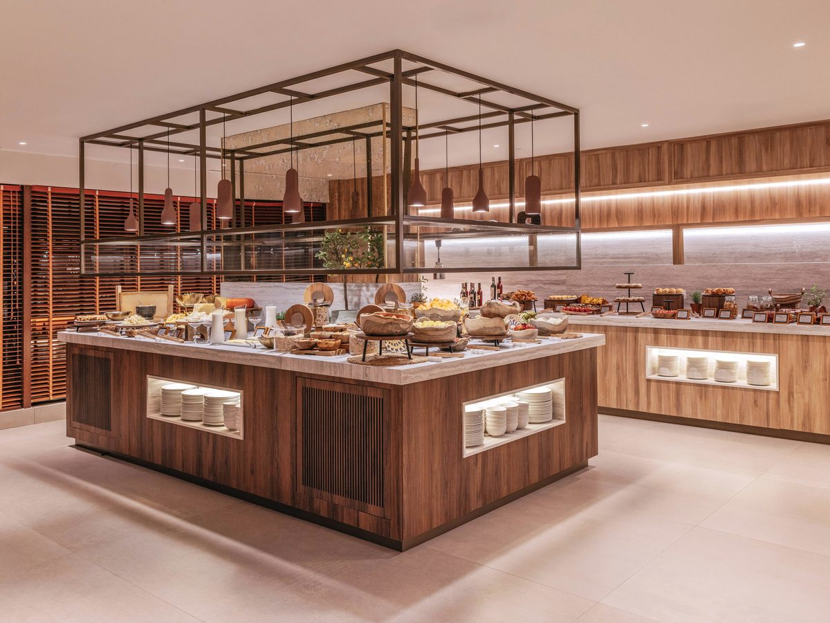 A long wooden buffet island laid with breads, pastries and hot dishes under warm lighting in the breakfast restaurant.