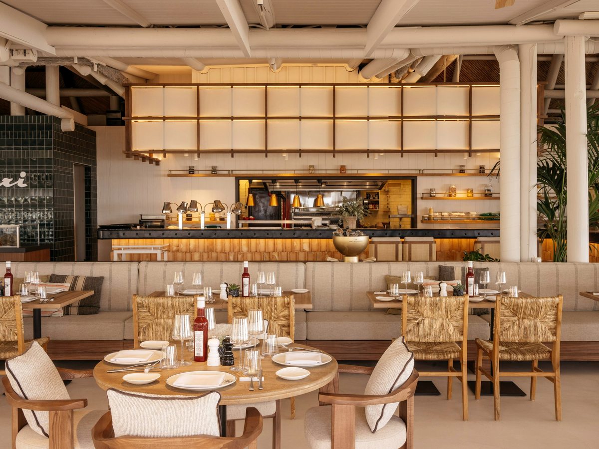 A bright restaurant with woven-back chairs and banquette seating facing an open kitchen behind a long counter.