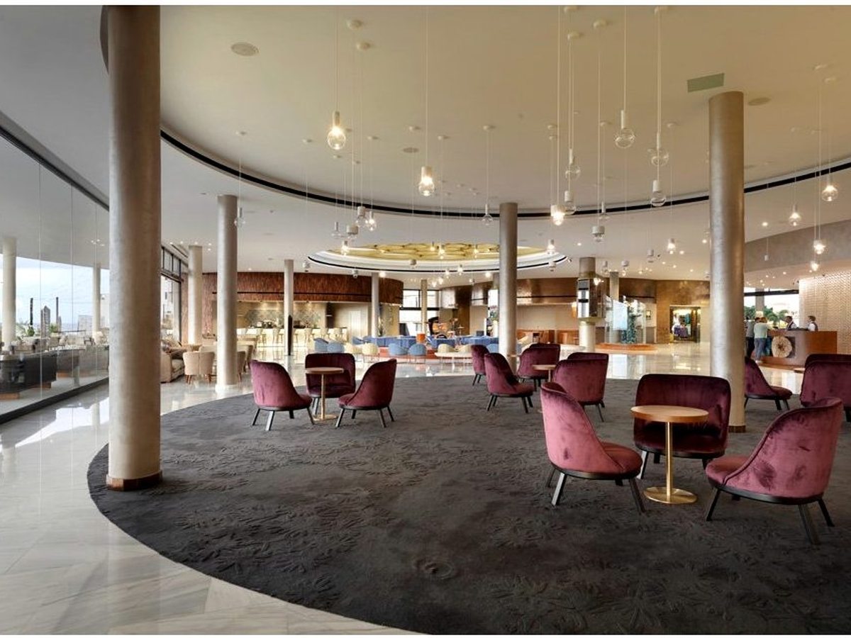 Circular lobby lounge with burgundy armchairs and round tables on a grey carpet, ringed by floor-to-ceiling windows and tall columns.