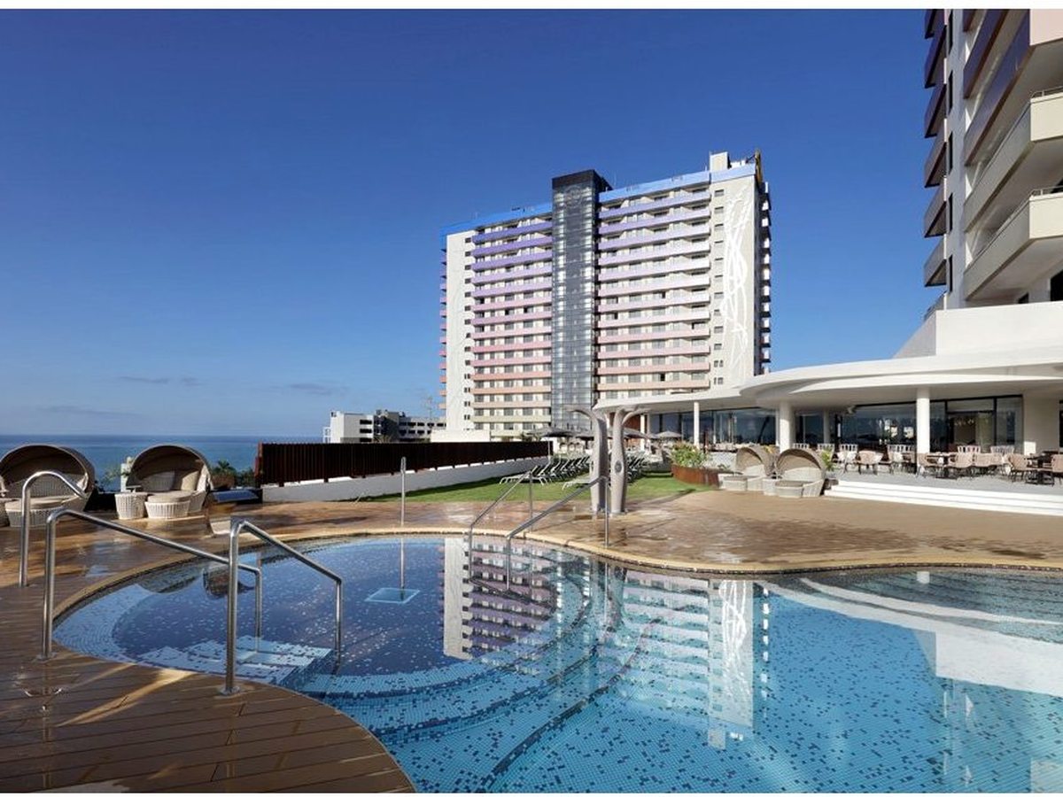 A curved pool with sun loungers along a wooden deck, the resort towers rising behind and the sea beyond.