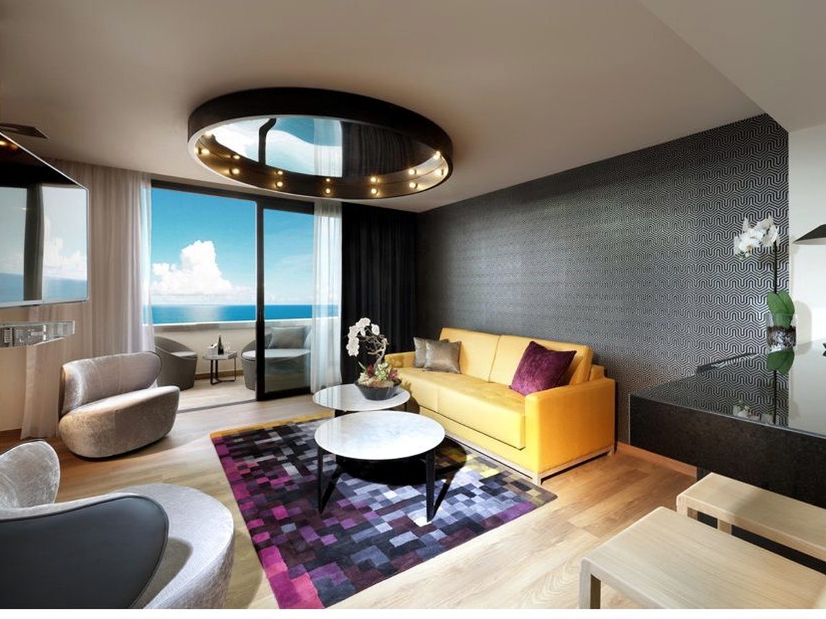 Suite living area with a yellow sofa, a round coffee table and lounge chairs beside sliding doors onto a sea-view balcony.