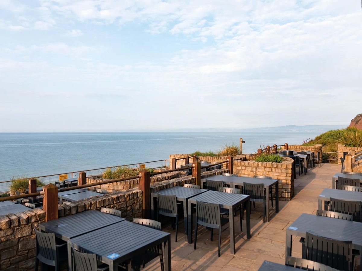 An outdoor dining terrace at Haven Devon Cliffs with rows of grey tables and chairs on stone paving behind a low wall, overlooking the sea and the coast beyond.