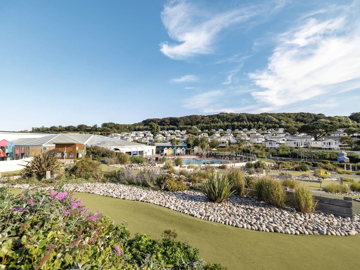 Elevated view over Haven Devon Cliffs: the outdoor pool complex and planted gardens in the foreground with rows of caravans rising up a wooded hillside behind, under a blue sky.