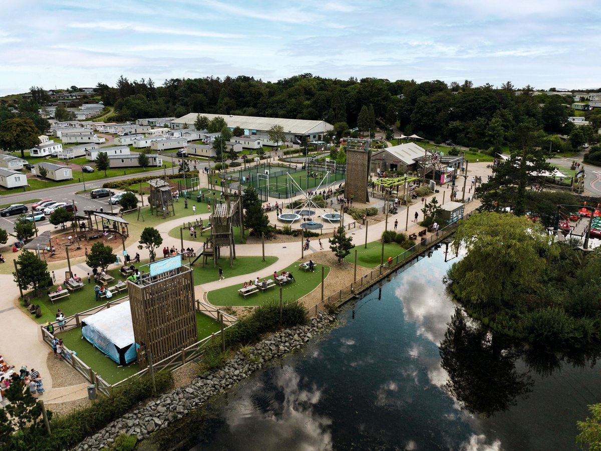 Aerial view of the park's adventure village with a climbing tower, sports courts, trampolines and adventure play beside a lake, with rows of caravans behind.