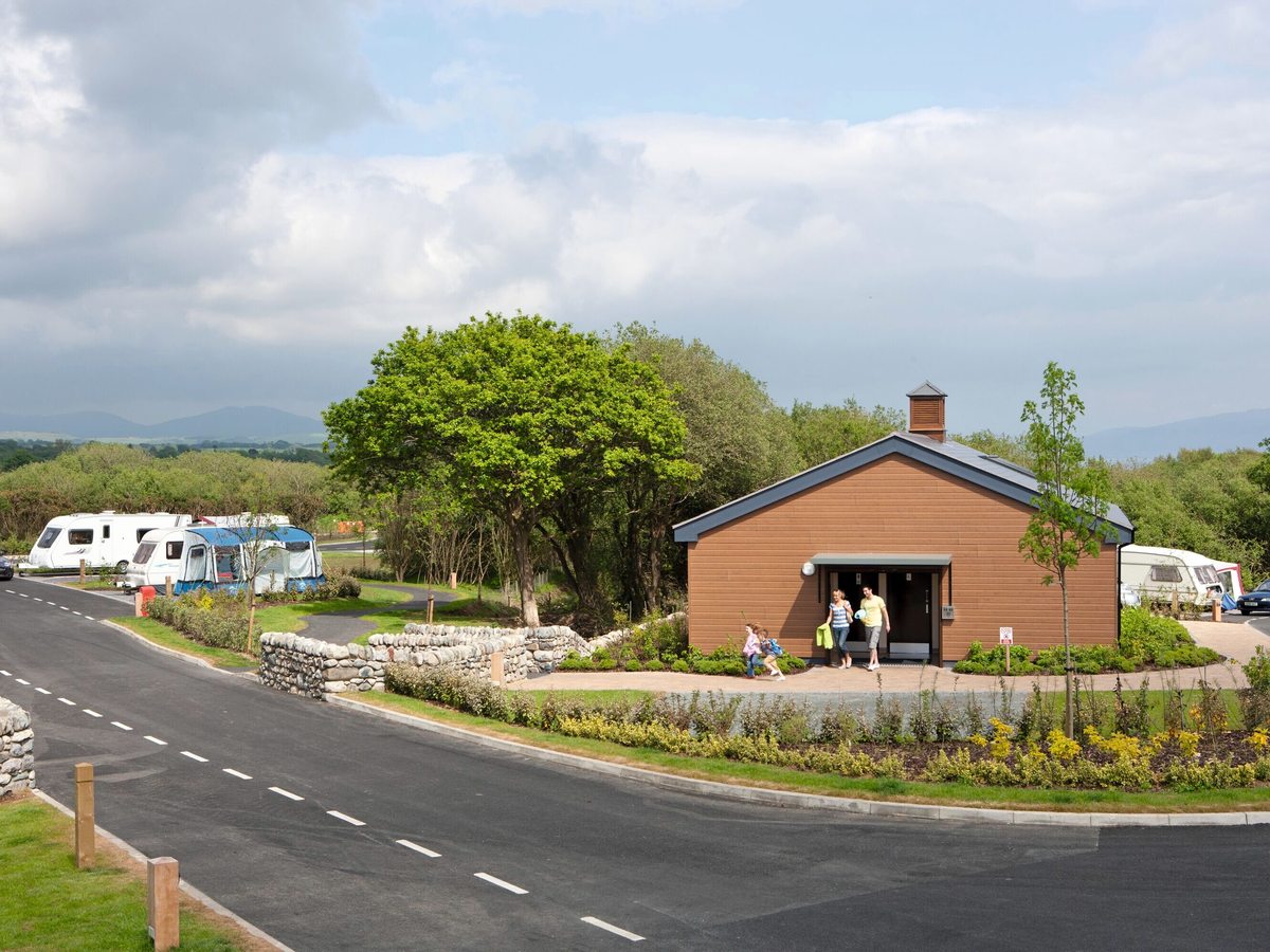 An internal park road curving past a timber-clad facilities building, landscaped beds and touring caravans, with wooded hills in the distance.