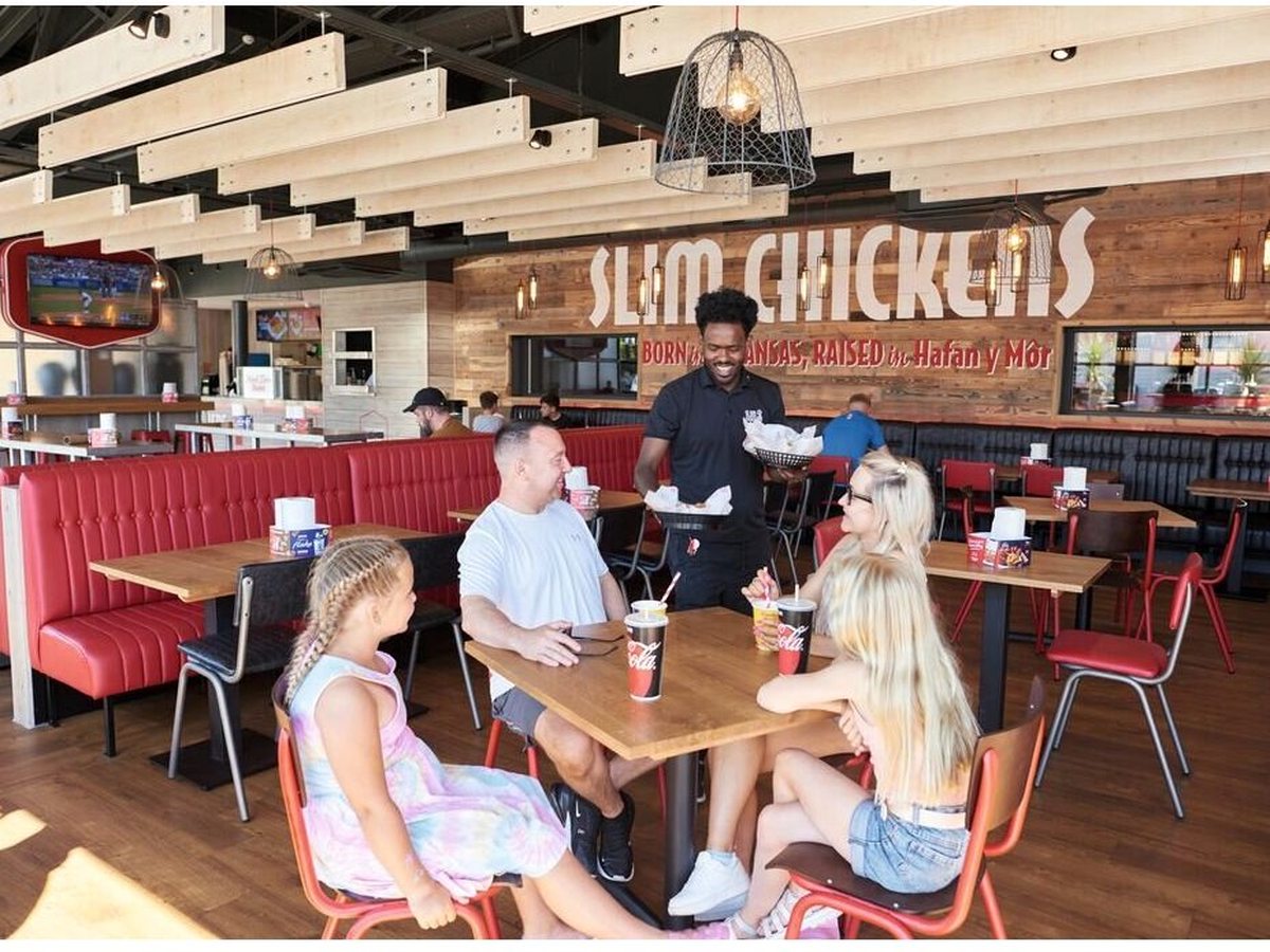 The interior of Slim Chickens with red booth seating, loose tables and a family eating at a table.