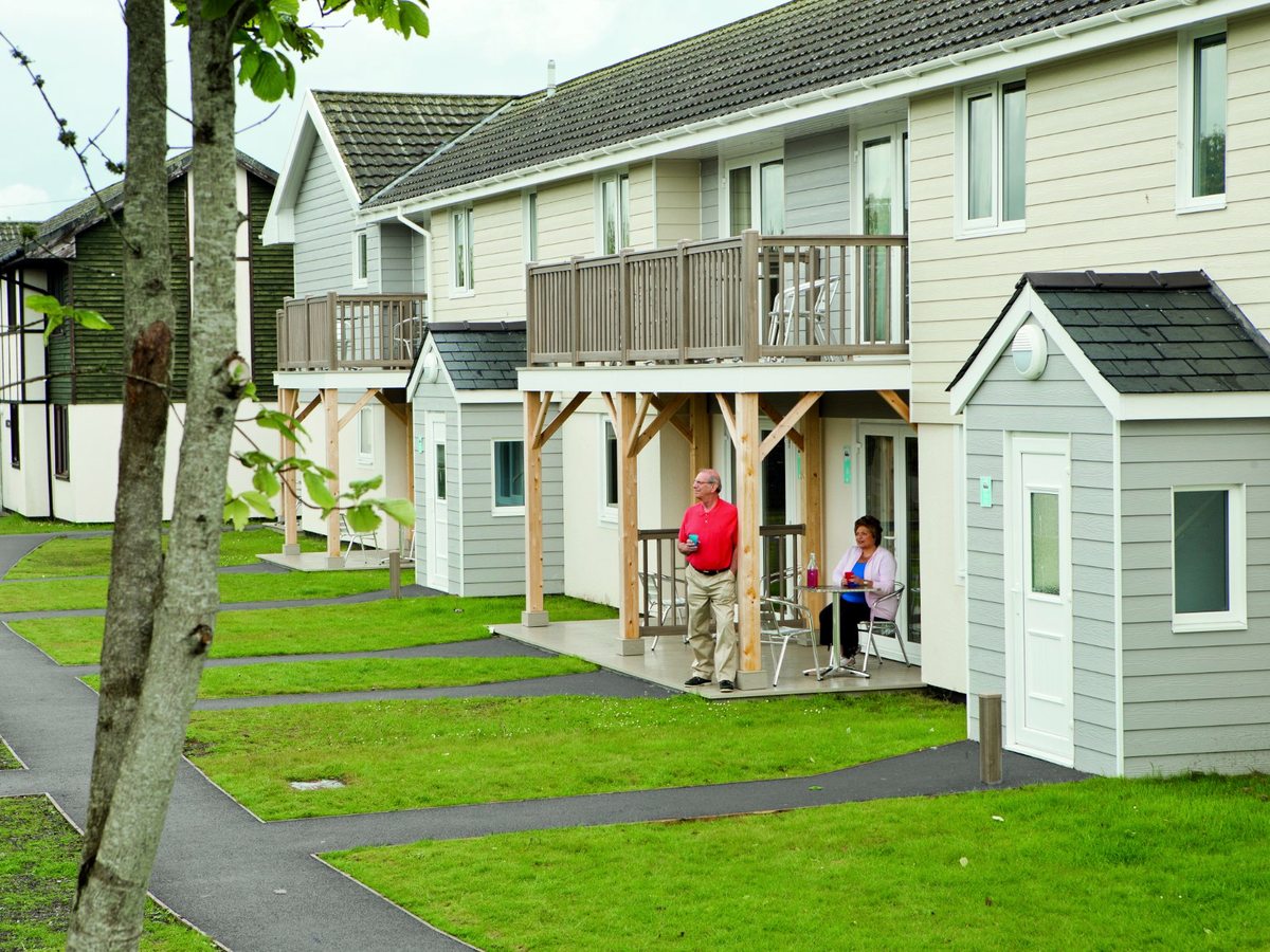 Two-storey self-catering apartment blocks with timber balconies and covered patios, reached by a paved path across a lawn.