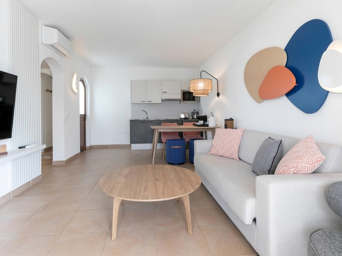An open-plan suite living room with a pale sofa, round wooden coffee table, a dining table with stools and a kitchenette with cabinets, sink, microwave and coffee machine, and abstract discs on the wall.