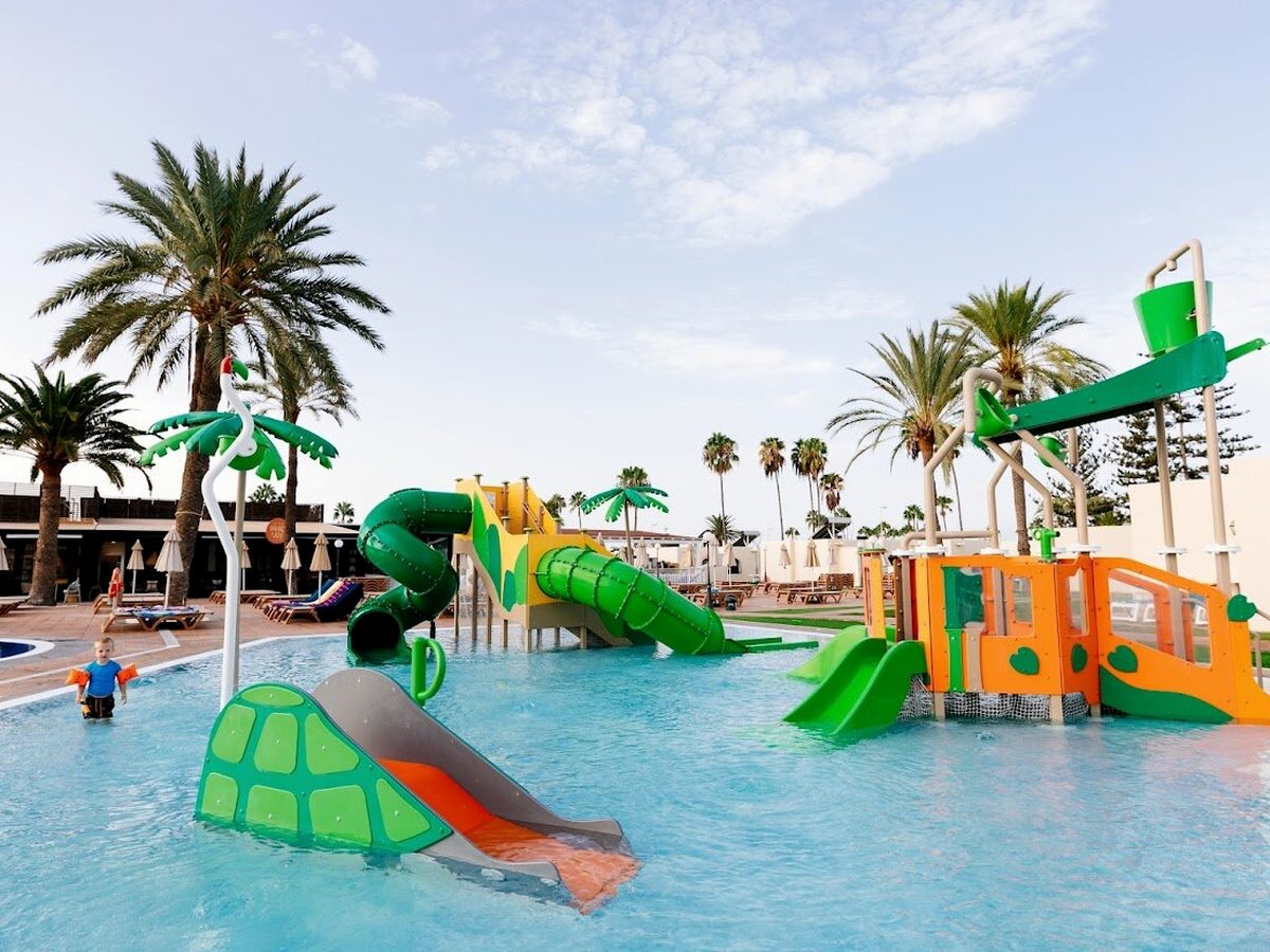 A shallow children's splash park with green palm-shaped and turtle slides, water features and small children playing, palms and blue sky behind.