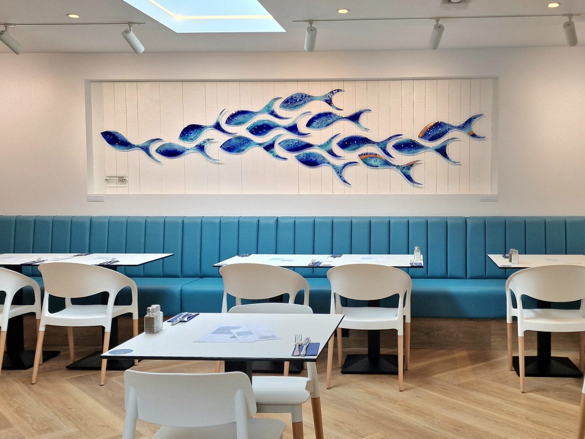 A bright restaurant with white chairs, a teal banquette and a blue shoal-of-fish artwork on a white plank wall.