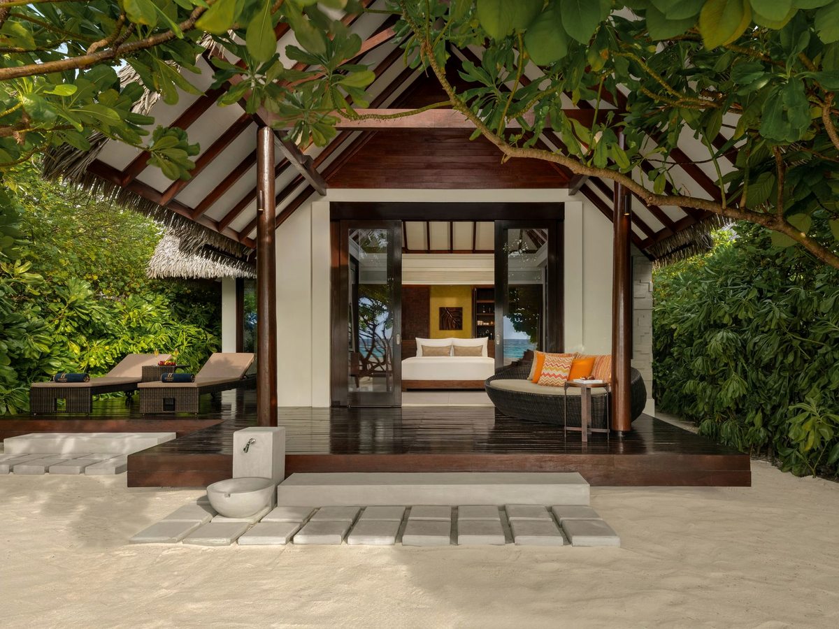 A thatched beach villa entrance with a covered terrace, daybed and loungers on a sandy path through tropical planting.
