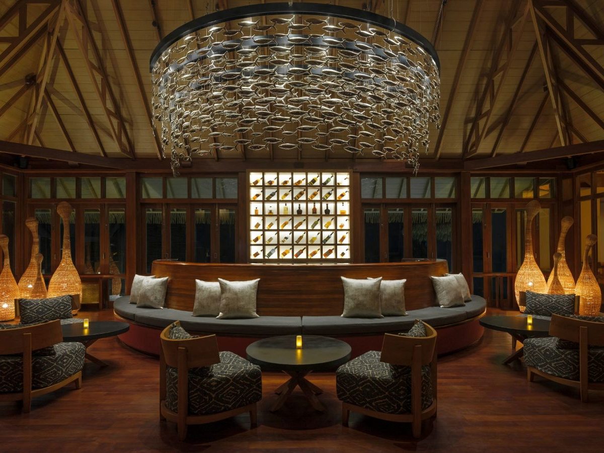 An open-sided lounge with a large woven pendant light, timber columns and cushioned sofas under a high thatched roof.
