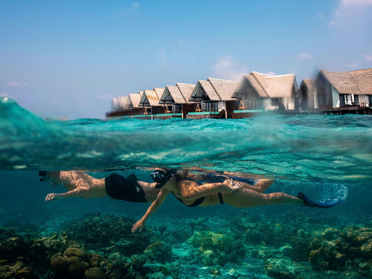 Two snorkellers floating over a coral reef in clear shallow water, with the overwater villas behind.