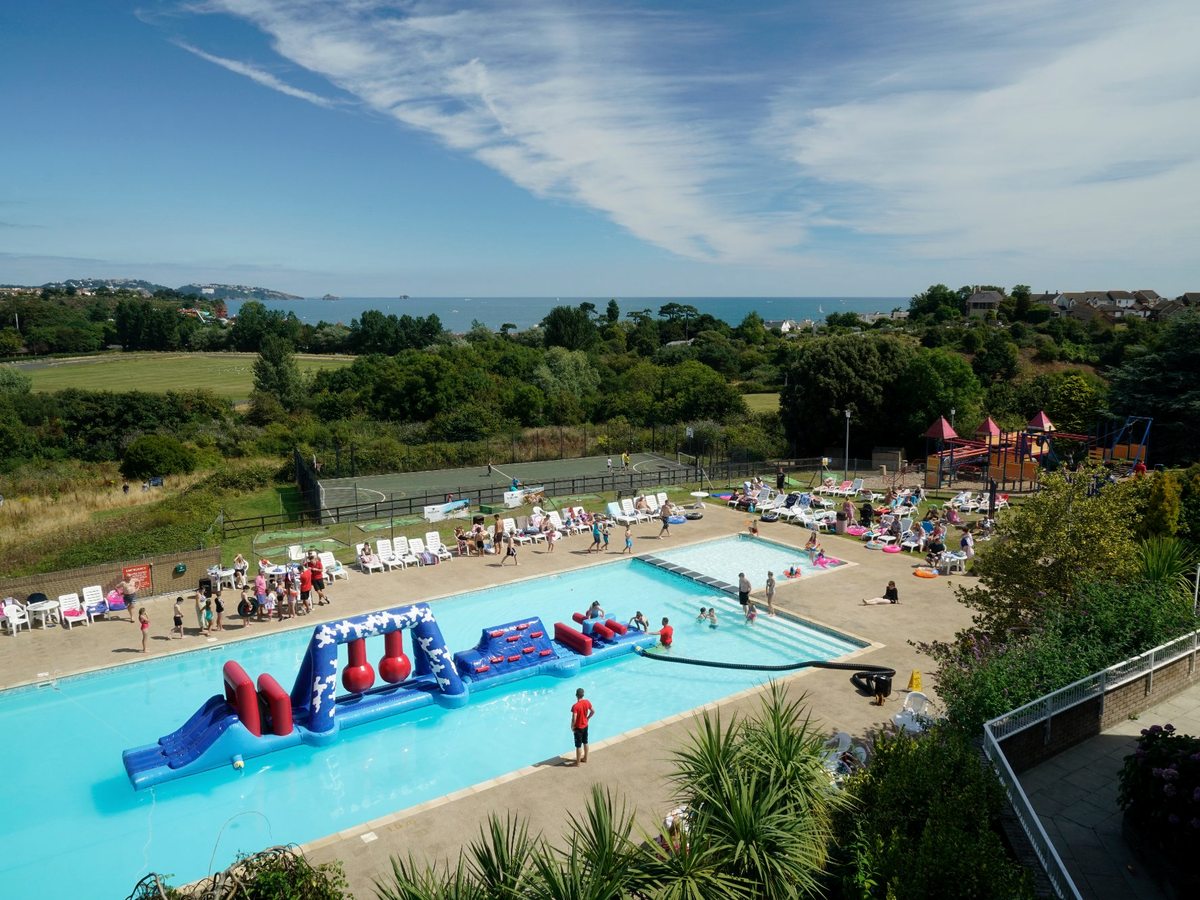 An aerial view of the outdoor pool with a large blue-and-red inflatable obstacle course, families around it and the sea beyond.