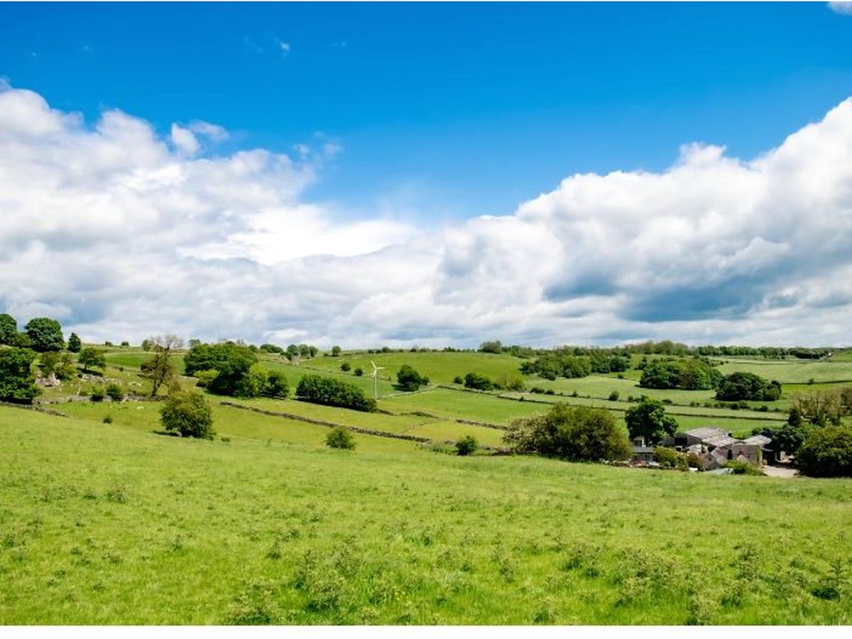 Rolling green fields bounded by dry-stone walls under a big sky, with a wind turbine and farm buildings in the distance, in the White Peak countryside around Hoe Grange.