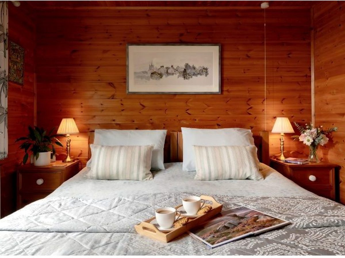 A log-cabin double bedroom with pine-clad walls, a bed made up in white linen with striped cushions, bedside lamps and a breakfast tray on the bed.