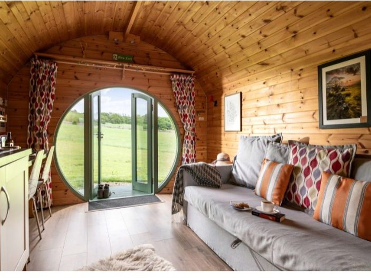 A pine-lined glamping pod interior with a grey sofa and scatter cushions, a small kitchen area and a round window with glazed doors opening onto green fields.