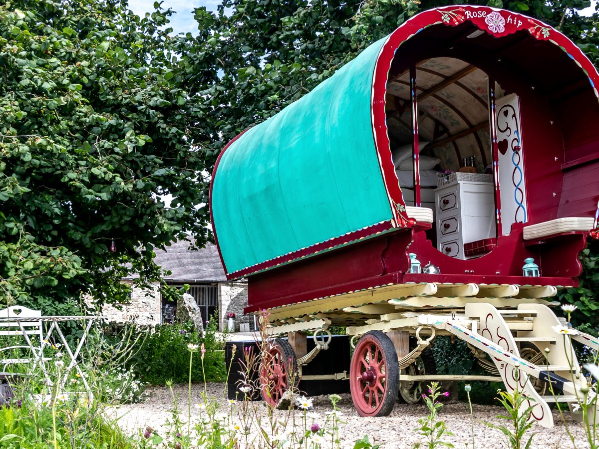 A traditional bow-top gypsy caravan painted deep red with a green canvas roof and red spoked wheels, standing among wildflowers by a stone barn.