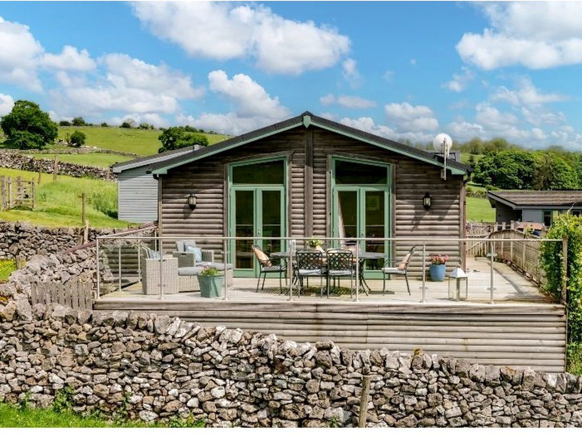 A single-storey grey-clad log cabin with green window frames and a decked terrace, set among green fields and dry-stone walls under a blue sky.