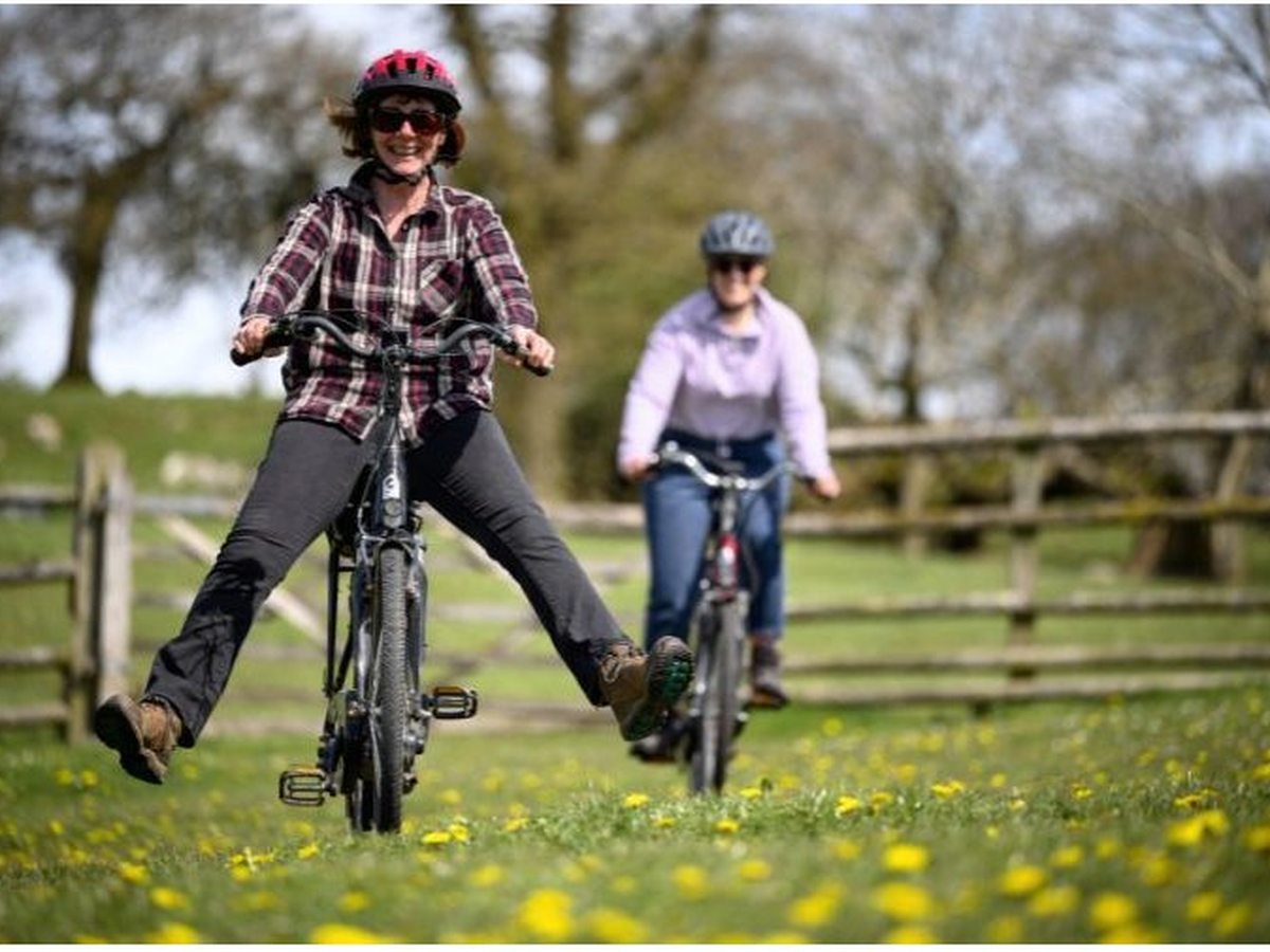 Two women in helmets riding bikes across a buttercup-strewn field by a wooden fence, one freewheeling with her legs out, both smiling.