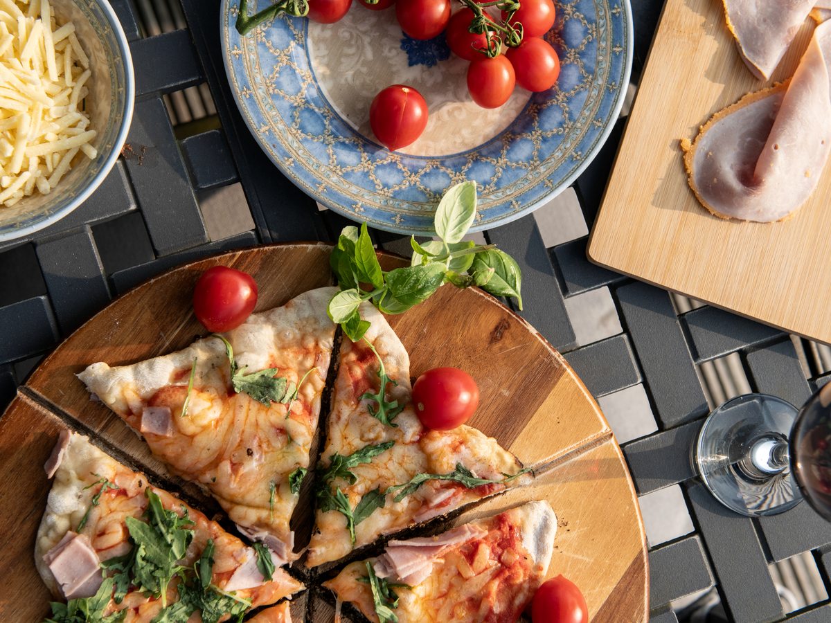 A thin-crust ham and rocket pizza on a wooden board on an outdoor table, with grated cheese, sliced ham, cherry tomatoes and a glass of red wine alongside.