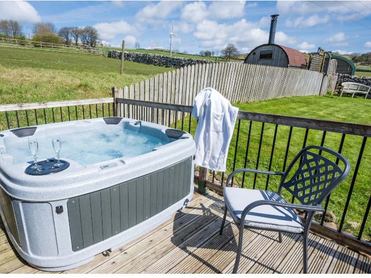 A bubbling hot tub with two glasses on its edge on a timber deck with metal railings, looking out over green fields and dry-stone walls, with a barrel sauna in the garden beyond.