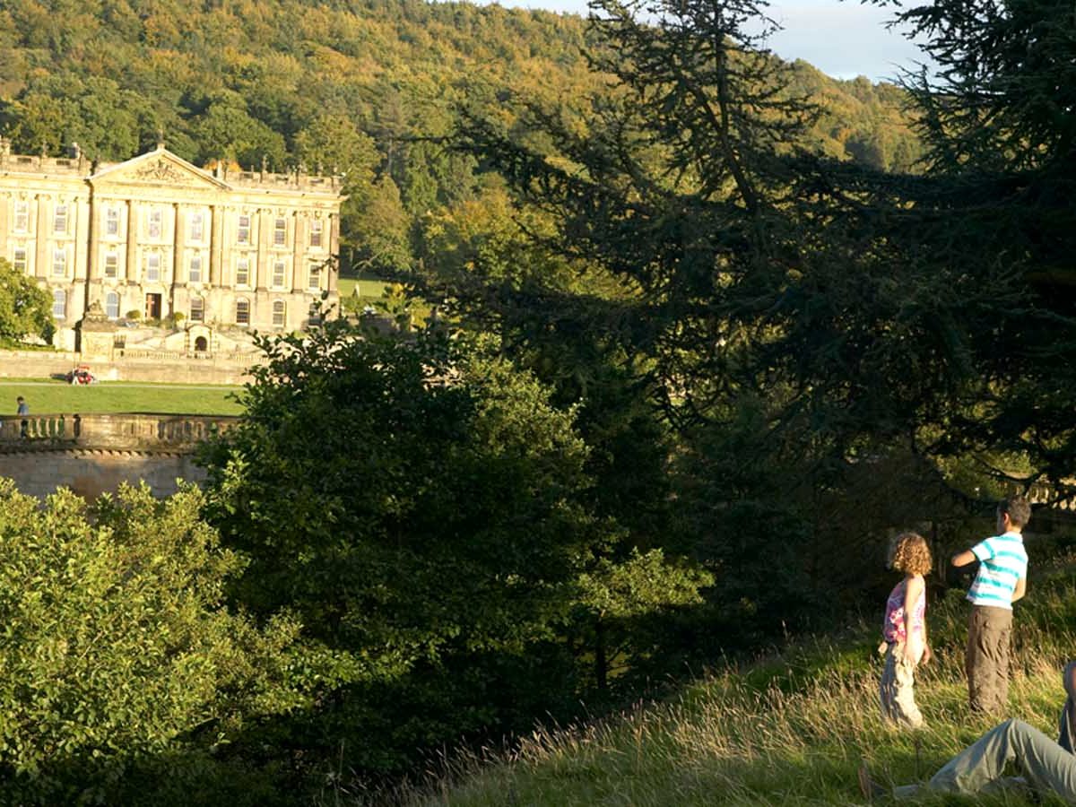 The honey-stone frontage of Chatsworth House across parkland, framed by trees, with two people looking towards it from a grassy slope.