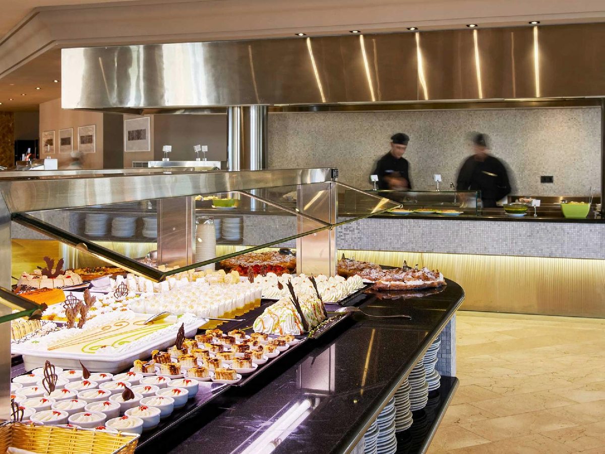 Long buffet counter laid with rows of individual cakes, tarts and mousses behind a glass guard, with chefs at a station behind.