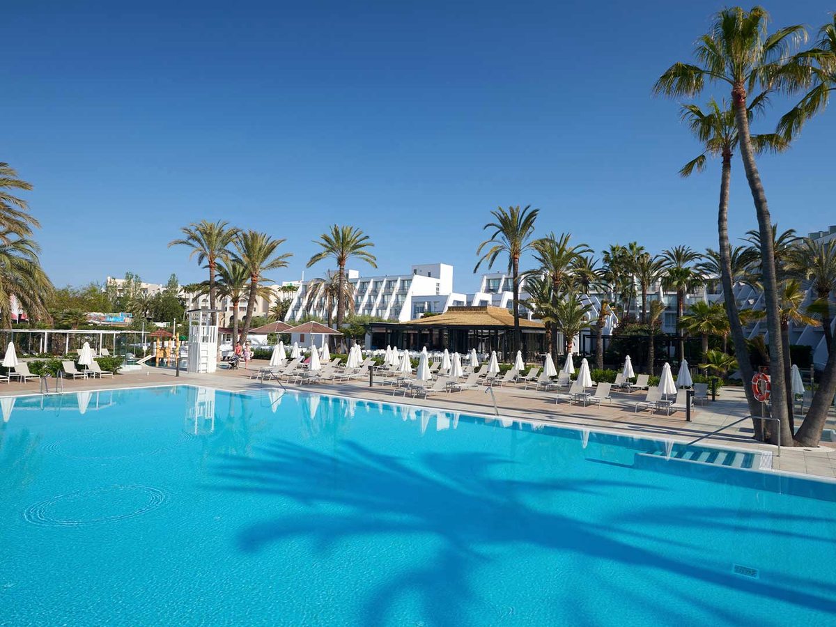 Large outdoor pool with a thatched pool bar and rows of loungers, the resort's white sawtooth-balconied blocks and tall palms behind.