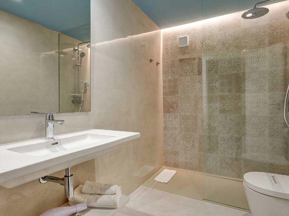 Bathroom with a long white washbasin, a large mirror and a walk-in shower behind a glass screen, with patterned Mediterranean wall tiles.