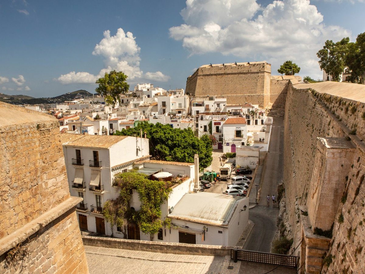 Whitewashed houses packed within the honey-stone walls of Dalt Vila, the old town climbing the hill with a bastion above and hills beyond.