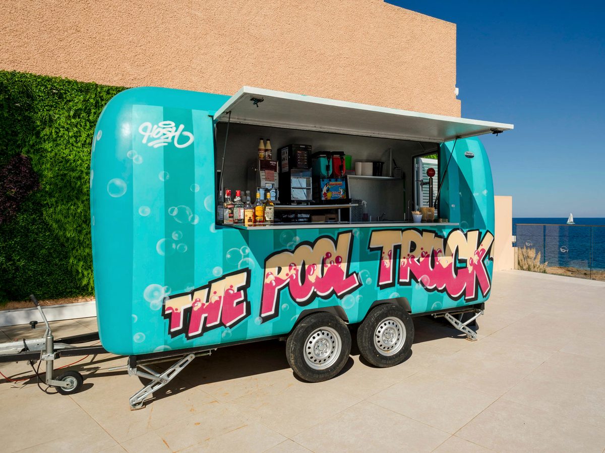 A turquoise converted trailer serving as a bar, its side shutter up to show a coffee machine and bottles, standing on a terrace by the sea.