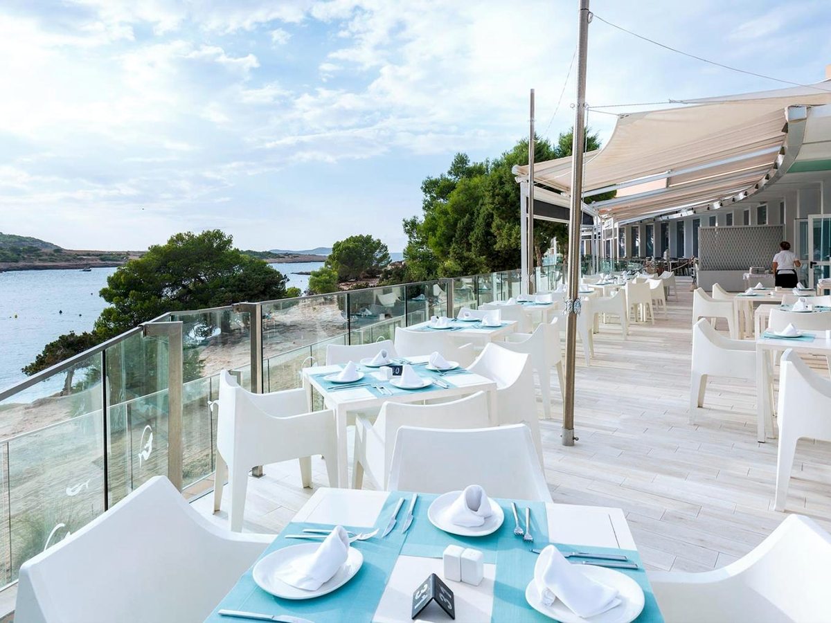White chairs and tables set with turquoise runners on a decked terrace overlooking the beach and bay, under a stretched white canopy.