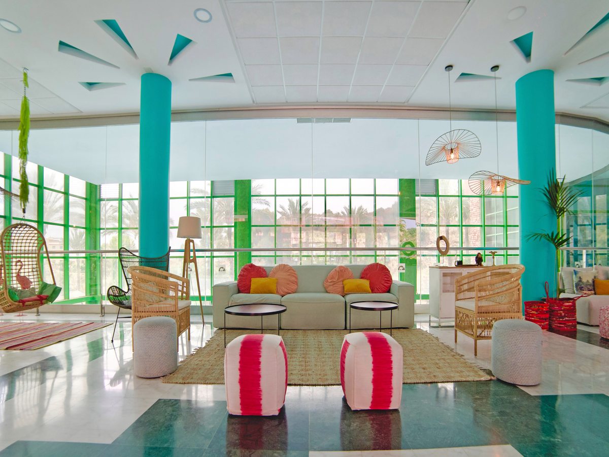 A bright glass-walled lounge with pastel sofas, cane armchairs and a hanging egg chair beside turquoise-clad columns and floor-to-ceiling windows.