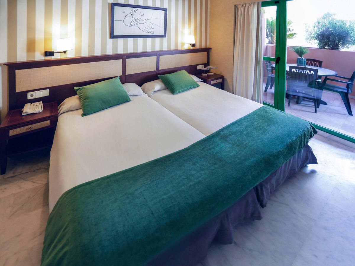 A twin room with two single beds under a shared wooden headboard, turquoise runner and cushions, a striped feature wall and a glazed door to a furnished balcony.