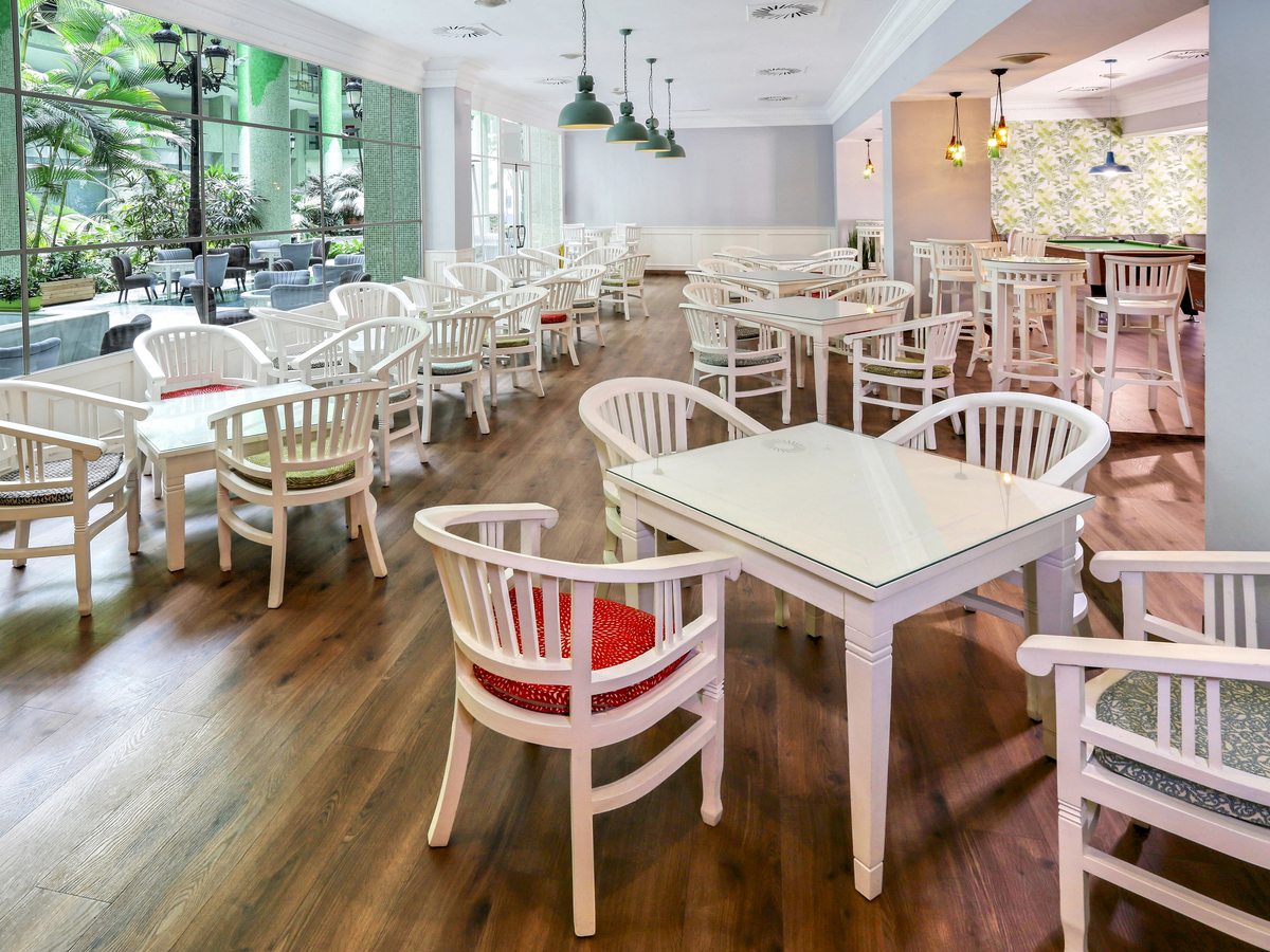 A restaurant dining room with white wooden chairs and tables on a wood floor beside large windows looking onto greenery.