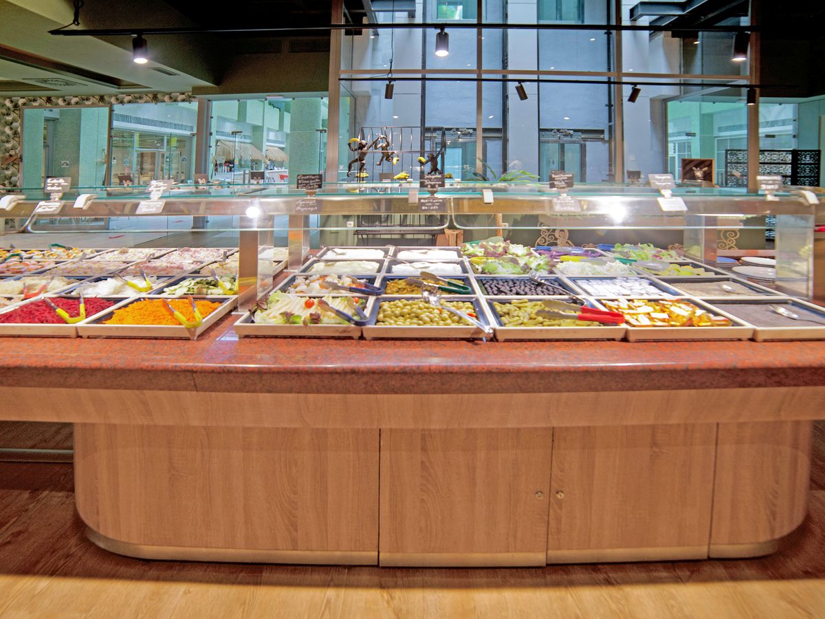 A long wooden buffet island laid with trays of salads, vegetables, olives and cold dishes under a glass guard, in front of tall windows.