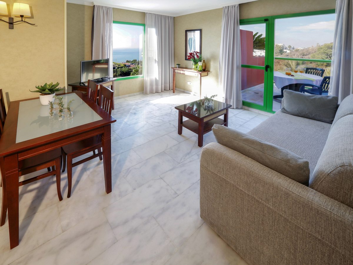A suite living and dining area with a grey sofa, wooden dining table and coffee table, with green-framed doors to a terrace and the sea beyond the window.