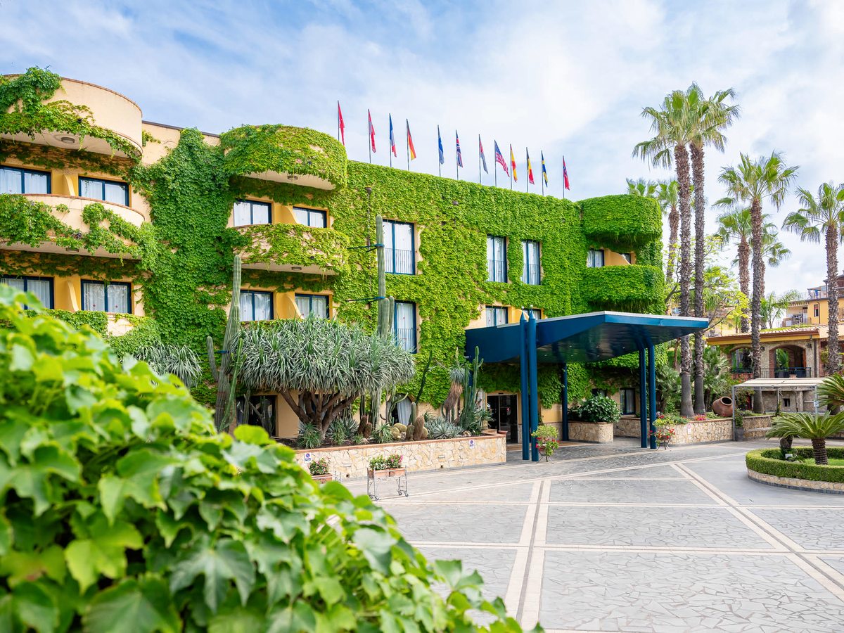 The four-storey Hotel Caesar Palace, its facade covered in green ivy, with a row of international flags along the roofline, tall palm trees and a covered entrance porch.