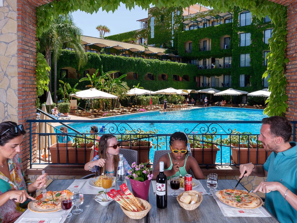A family eating pizza and pasta at a table on a covered terrace, with the hotel pool, cream parasols and the ivy-covered building behind.