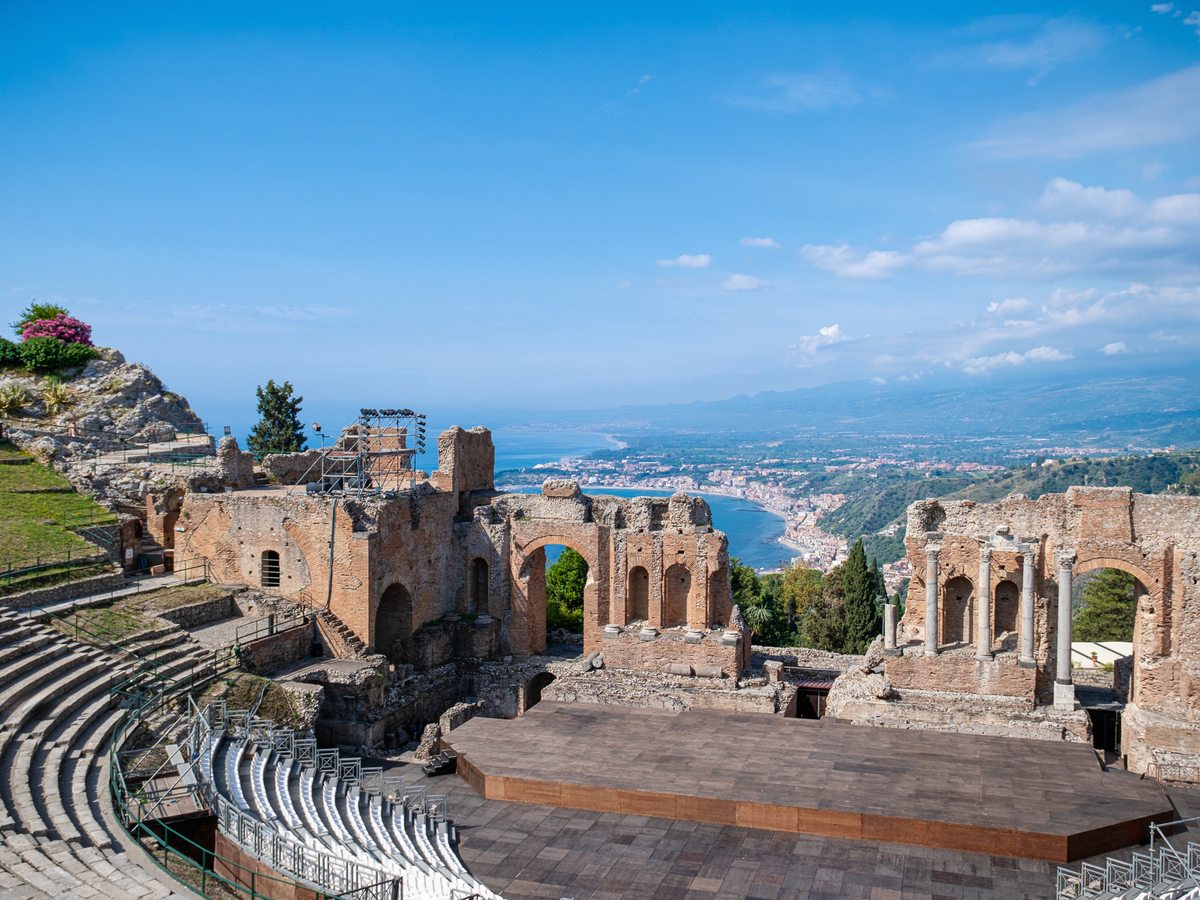 The stone ruins and tiered seating of the ancient Greek theatre at Taormina, looking out over the bay and coastline far below under a blue sky.