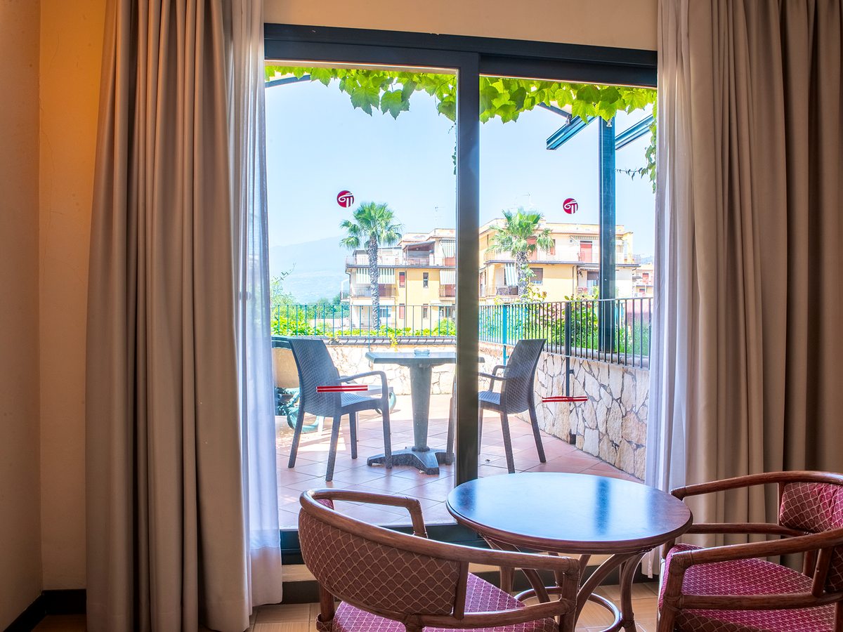 A hotel room with rattan chairs and a table by full-length glass doors opening onto a private terrace with its own table and chairs, looking out to houses, palms and hills.