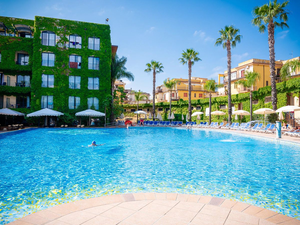 A large outdoor pool with blue sun loungers and cream parasols along one side, tall palm trees and the ivy-covered hotel behind under a clear blue sky.